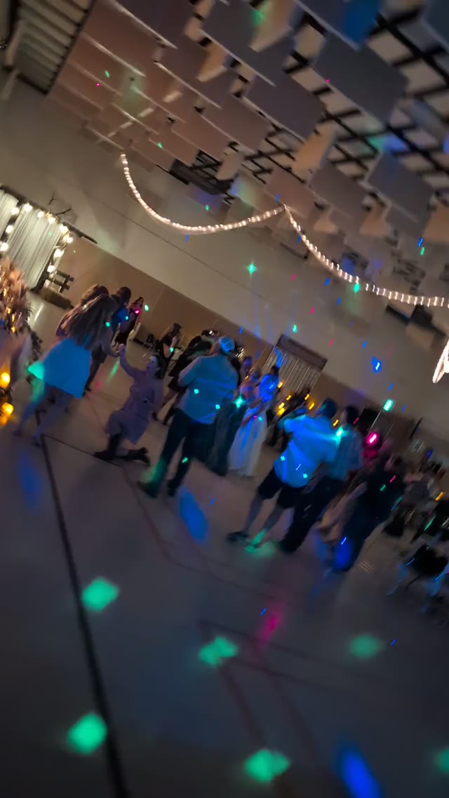 A packed dance floor in a community hall under string lights, with disco spots sweeping the crowd