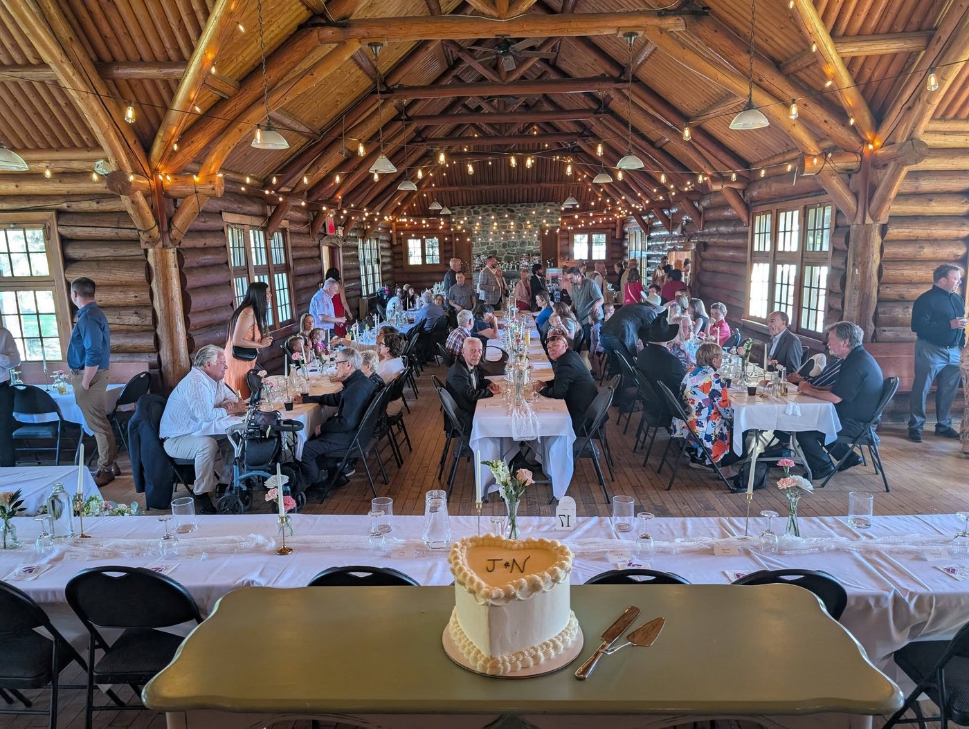 A real DJ Fetchy wedding reception — guests seated in a log hall strung with lights, with the couple's 'J+N' cake on the head table